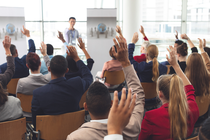 Rear view of diverse business people raising hands while they are sitting in front of Asian businessman at business seminar in office building.