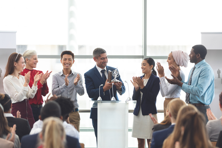 Front view of mixed race businessman holding award on podium with colleagues in front of business professionals at business seminar in office building.