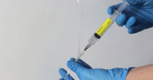 ALT: Provider with blue gloves holding syringe of Ketamine and injecting into clear tube