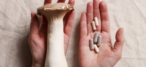 A large mushroom is held in one hand beside an open hand holding several white capsules and pills, set against a light-colored fabric background.