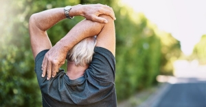 Back view of an older man stretching his arms behind his head while outdoors.