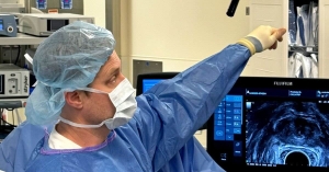Dr. Sharpel in surgical scrubs and a mask, pointing toward an ultrasound monitor displaying a prostate scan, set against a background of hospital equipment.