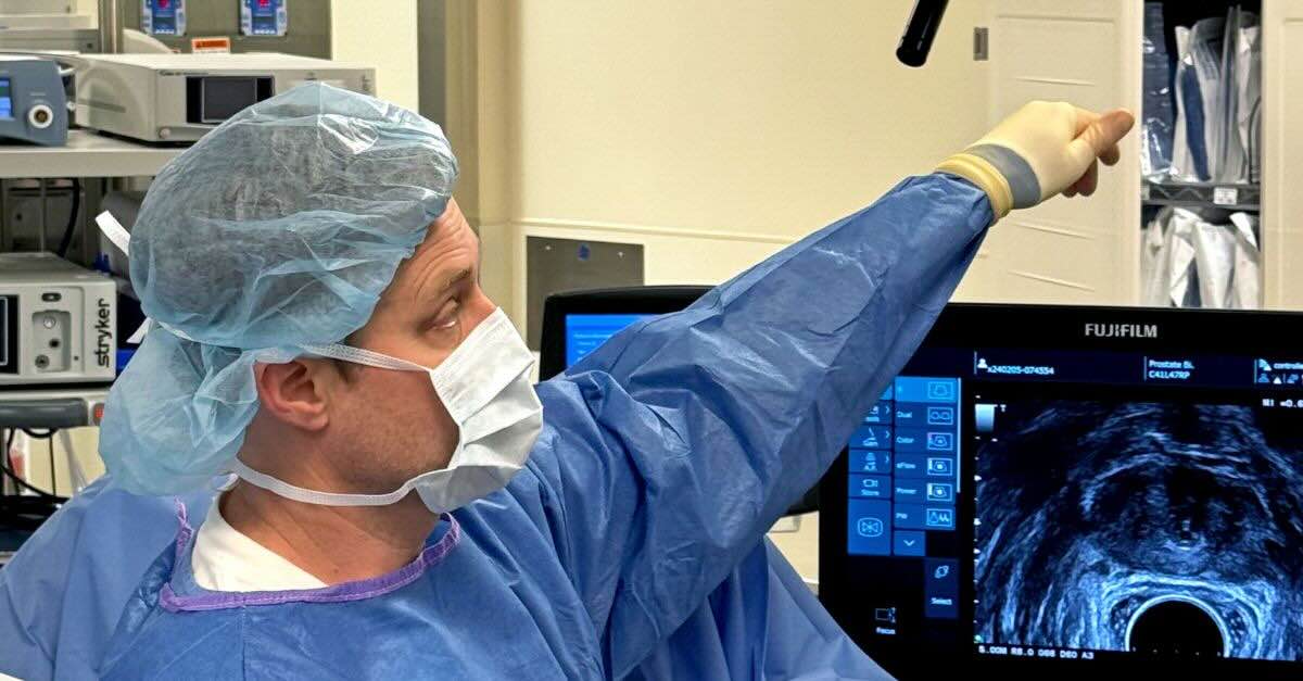 Dr. Sharpel in surgical scrubs and a mask, pointing toward an ultrasound monitor displaying a prostate scan, set against a background of hospital equipment.