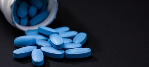 An open white prescription bottle lying on its side on a dark surface, with blue oval-shaped pills spilling out of the bottle opening and scattering across the dark background.