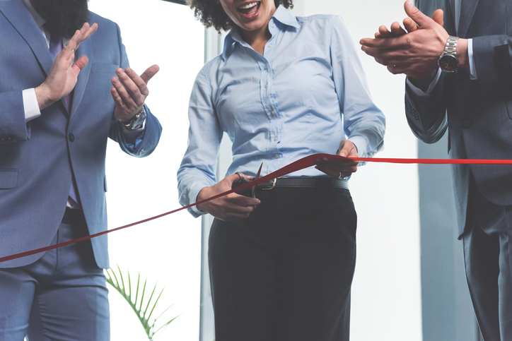 Team of ambitious young businesspeople on a ceremonial grand opening cutting red ribbon and clapping hands.