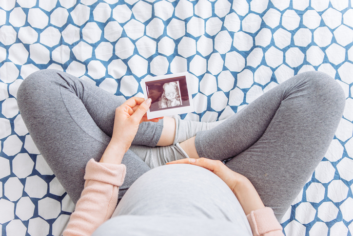 9 months pregnant young woman holding hand on her belly, she is sitting on bed and hold ultrasound image of baby, wondering about the urological risks of pregnancy.