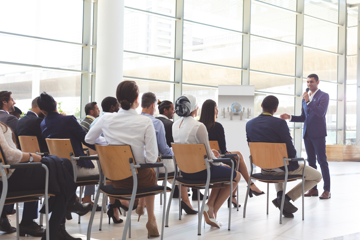 Front view of handsome mixed-race businessman speaking at business seminar with diverse business people listening to him at conference.