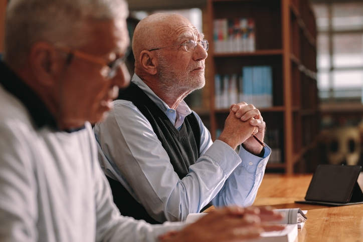 Two senior men sitting in classroom listening to a lecture. Elderly men sitting in a classroom with books and a tablet pc on the table.