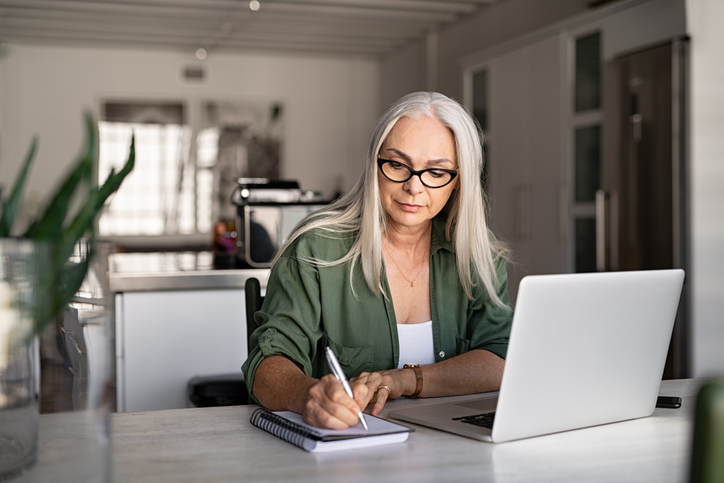 Senior stylish woman taking notes in notebook while using laptop at home. Old freelancer writing details on book while working on laptop in living room. Focused cool lady writing notary in notepad.