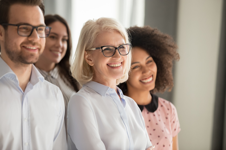 Side view of smiling diverse employees stand in row together posing for group picture in office, happy confident multiethnic team look at camera making photo, showing unity. Teamwork concept