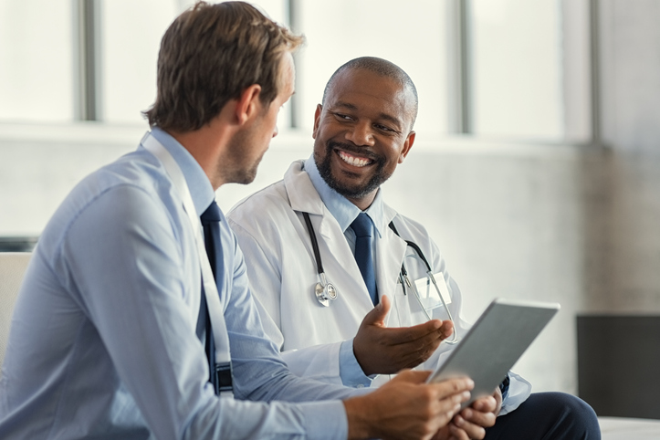 Two mature smiling doctors having discussion about patient diagnosis, holding digital tablet. Successful african surgeon discussing case after positive result. Representative pharmaceutical discussing with african happy doctor about new medicine.