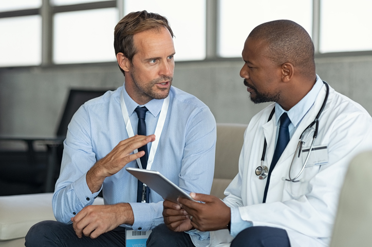 Pharmaceutical representative talking with doctor sitting on couch. African mature practitioner discussing results of the analysis with specialist while consulting diagnosis on digital tablet. Doctor in conversation with medical advisor in hospital room discussing patients report case.