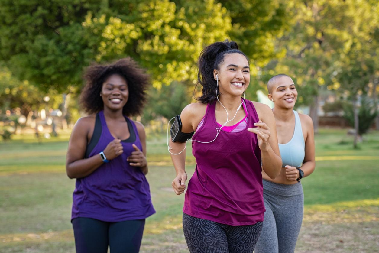 Group of curvy girls friends jogging together at park. Beautiful smiling young women running at the park on a sunny day. Female runners listening to music while jogging.